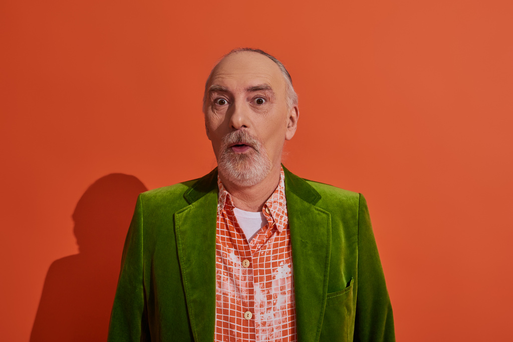 surprised and impressed senior man with bulging eyes looking at camera on red orange background, grey hair and beard, green velour blazer and trendy shirt, personal style and positive aging concept - Photo, Image