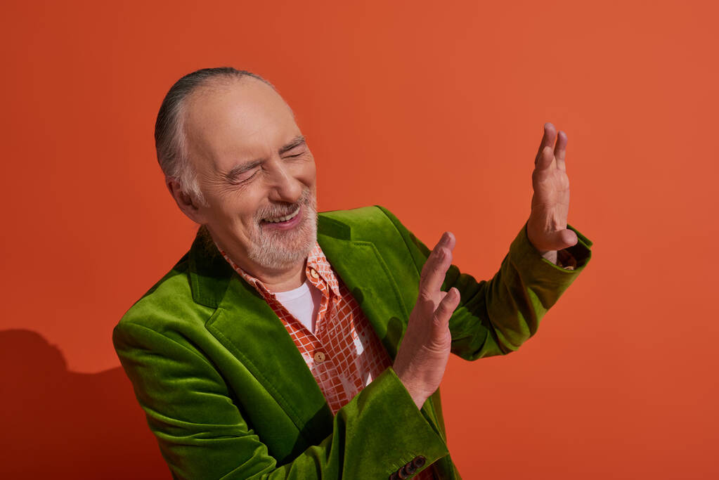 excited grey haired and bearded senior man in green velour blazer showing stop gesture and laughing with closed eyes on red orange background, personal style, fashionable aging concept - Photo, Image