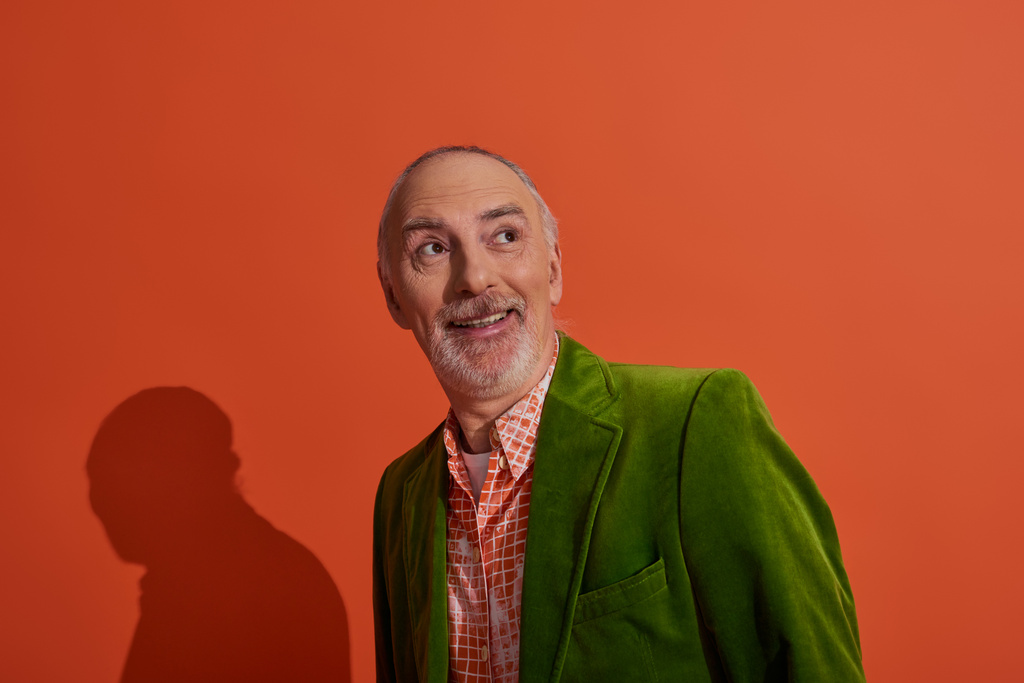 joyful and surprised senior man with grey hair and beard smiling and looking away on red orange background, fashionable casual clothes, green velour blazer, trendy shirt, positive aging concept - Photo, Image