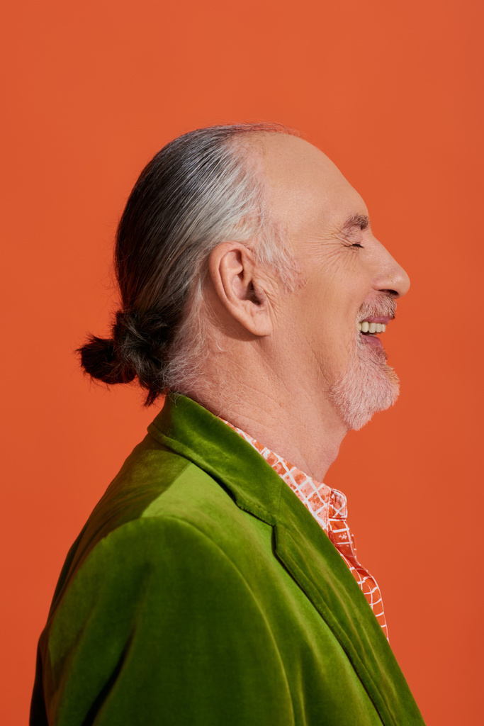 πλαϊνή άποψη του ενθουσιασμένοι και μοντέρνα παλαιότερο μοντέλο γέλιο με κλειστά μάτια σε ζωντανή πορτοκαλί φόντο, πορτρέτο του χαρούμενα και γενειοφόρος ηλικιωμένος άνδρας σε πράσινο βελούδινο σακάκι - Φωτογραφία, εικόνα