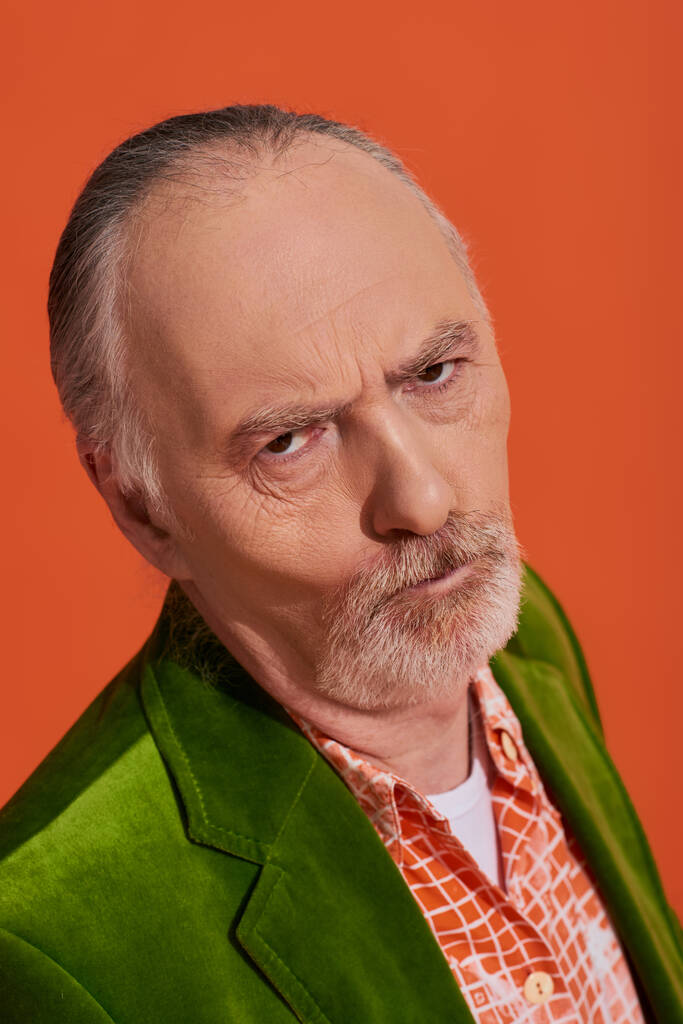 Porträt eines beleidigten älteren Mannes mit missmutigem Gesichtsausdruck, der auf leuchtend orangefarbenem Hintergrund in die Kamera blickt, älteres Modell, graue Haare, bärtiger, grüner Velours-Blazer, modisches Alterskonzept - Foto, Bild