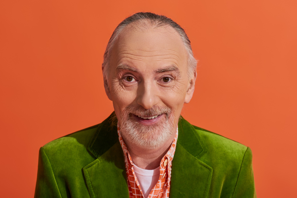 Porträt eines charismatischen älteren männlichen Models mit grauen Haaren, Bart und strahlendem Lächeln, das in die Kamera auf leuchtend orangefarbenem Hintergrund blickt, grünem Velours-Blazer, modischer Freizeitkleidung, positiver Alterung - Foto, Bild