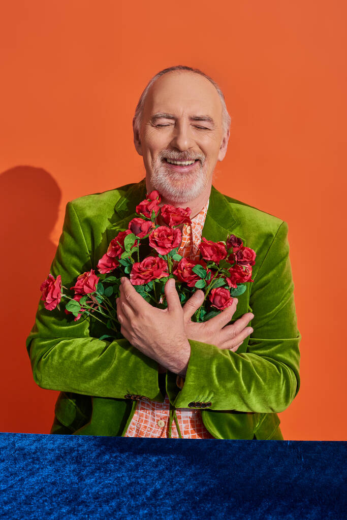 счастливый пожилой человек в зеленом бархатном блейзере, обнимающий красные розы и улыбающийся с закрытыми глазами возле стола с голубой велюровой тканью на ярком оранжевом фоне, позитивная концепция старения - Фото, изображение