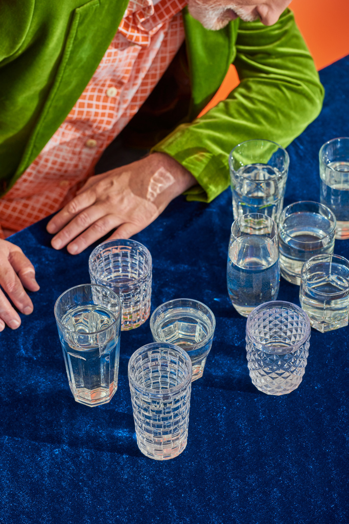 vista recortada de hombre mayor en blazer de terciopelo de moda y verde sentado cerca de vasos de cristal con agua clara en la mesa con tela de terciopelo azul sobre fondo naranja, simbolismo, concepto de plenitud de vida - Foto, imagen