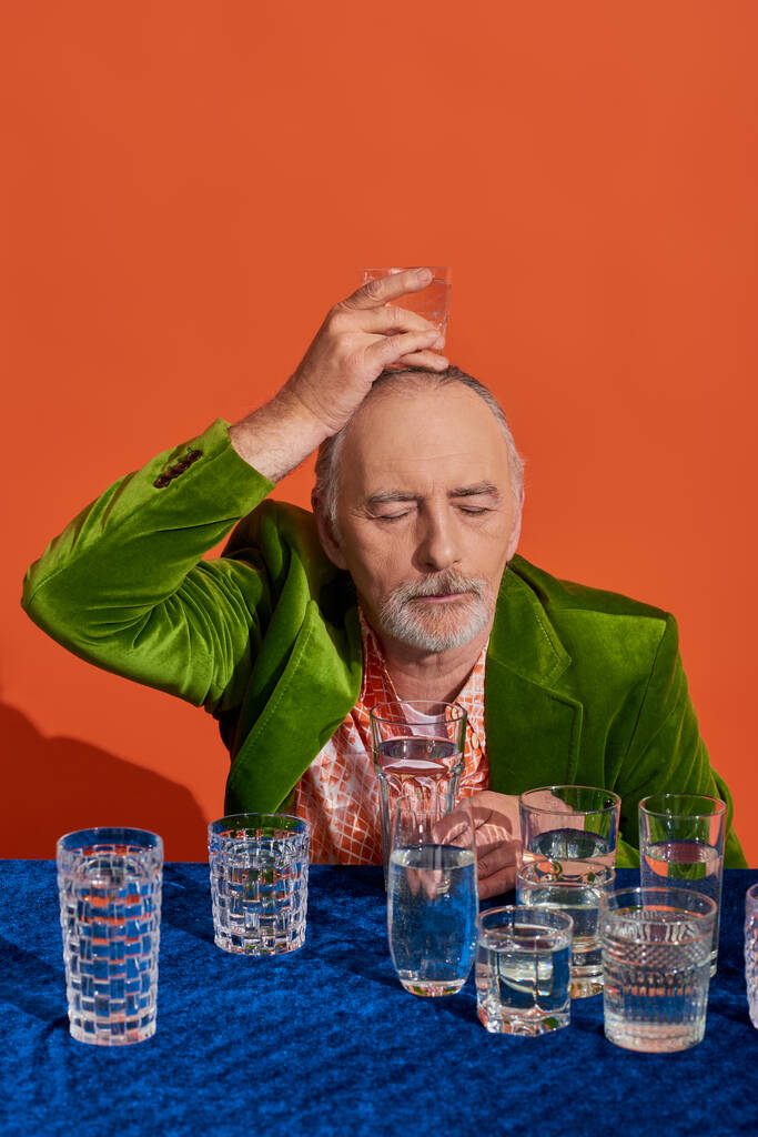 bärtiger älterer Herr mit geschlossenen Augen, in stylischem und grünem Samtblazer, hält ein Glas Wasser über dem Kopf auf orangefarbenem Hintergrund, alternde Bevölkerung, Symbolik, Konzept der Lebensfülle - Foto, Bild