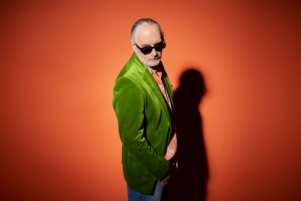 idősebb férfi modell elegáns alkalmi ruhák nézi kamera piros és narancssárga háttér árnyék, zöld velúr blézer, sötét napszemüveg, kifejező egyéniség, divatos öregedés koncepció - Fotó, kép