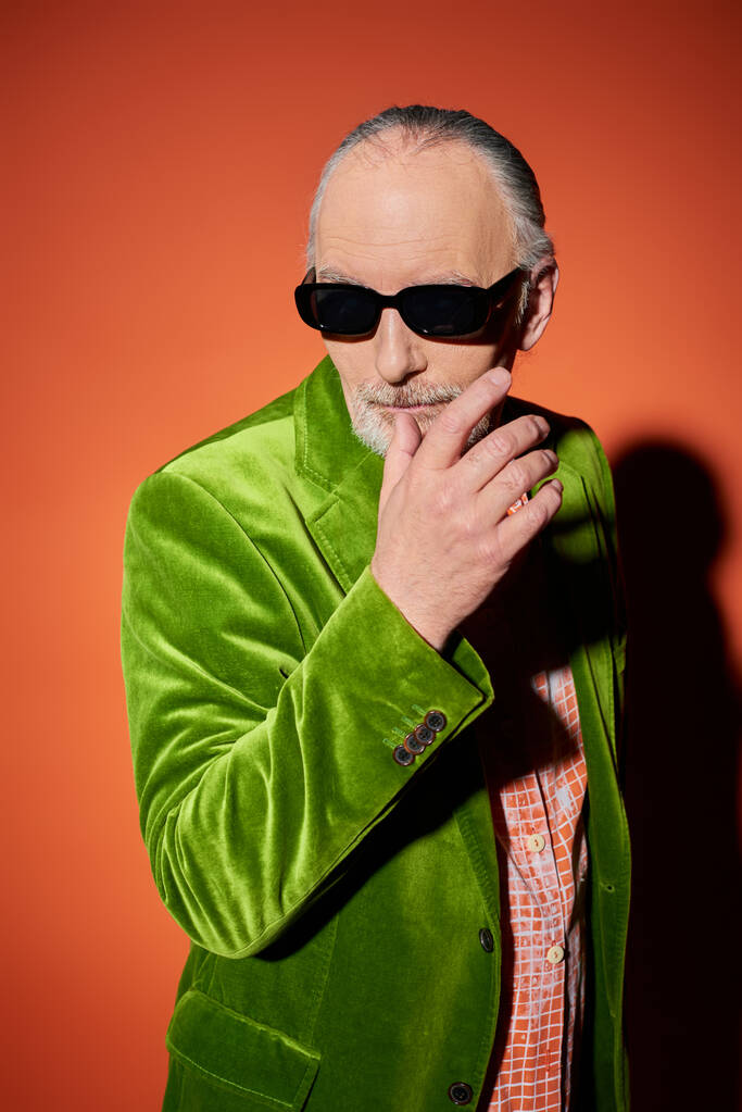 trendiger älterer Herr mit dunkler Sonnenbrille und grünem Velours-Blazer, der mit händennahem Bart posiert und vor rotem und orangefarbenem Hintergrund mit Schatten, Mode und Alterskonzept wegschaut - Foto, Bild