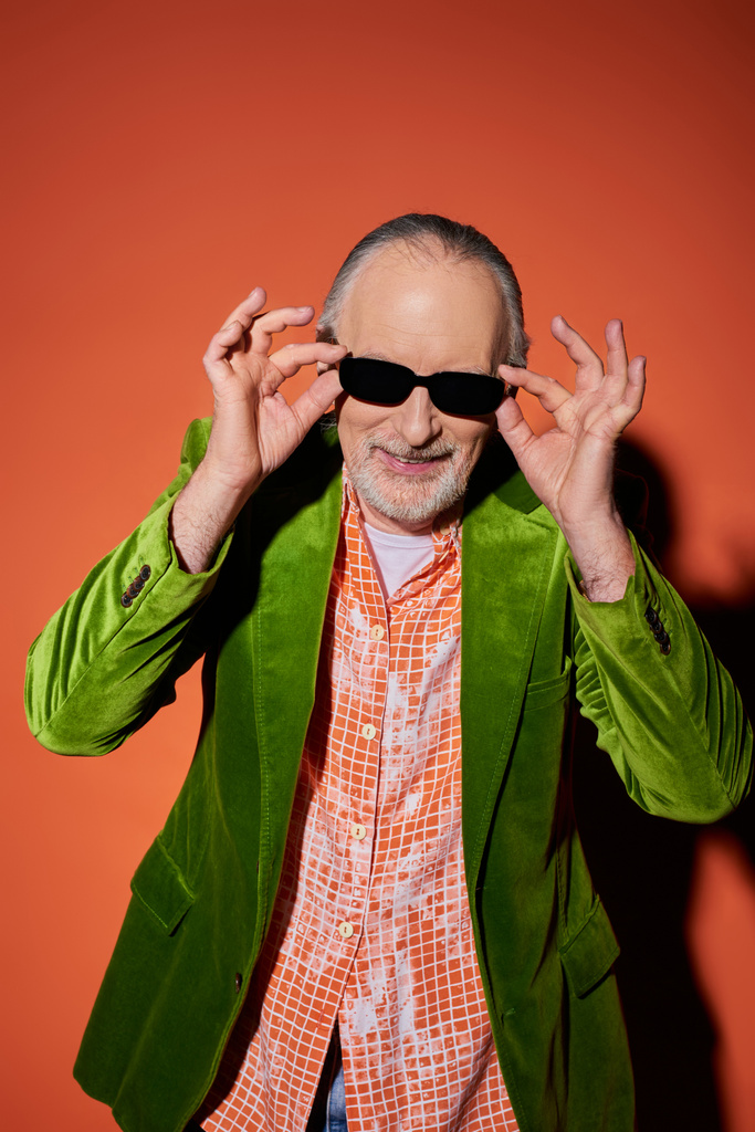 optimista idős férfi ápolt szakáll beállítása sötét napszemüveg és mosolygós kamera piros és narancssárga háttér, divat megjelenés, zöld velúr blézer, boldog és elegáns öregedés koncepció - Fotó, kép