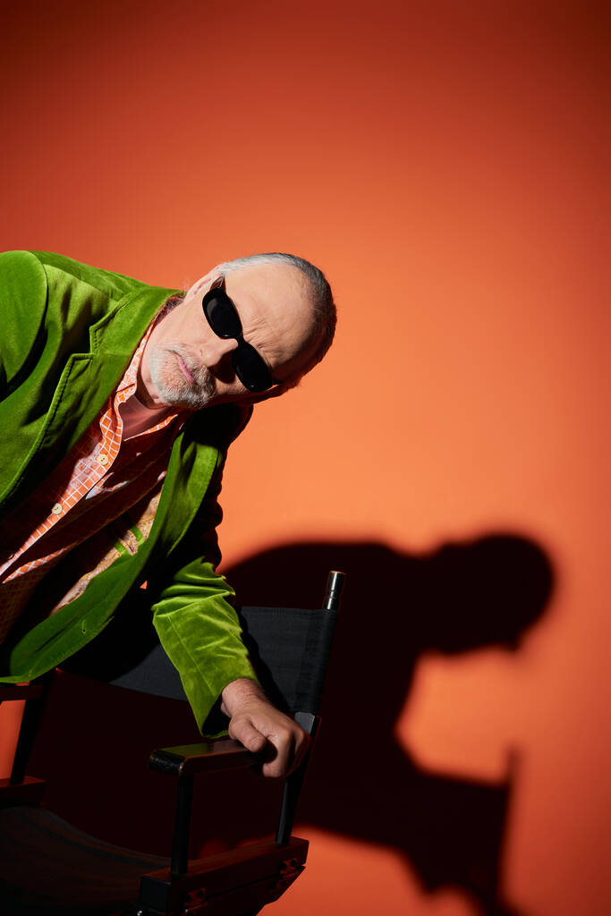 átgondolt és kíváncsi idős férfi sötét napszemüvegben és zöld velúr blézer áll közel a székhez, és félrenéz a piros és narancssárga háttér árnyékos, divatos öregedés, stílusos életmód - Fotó, kép