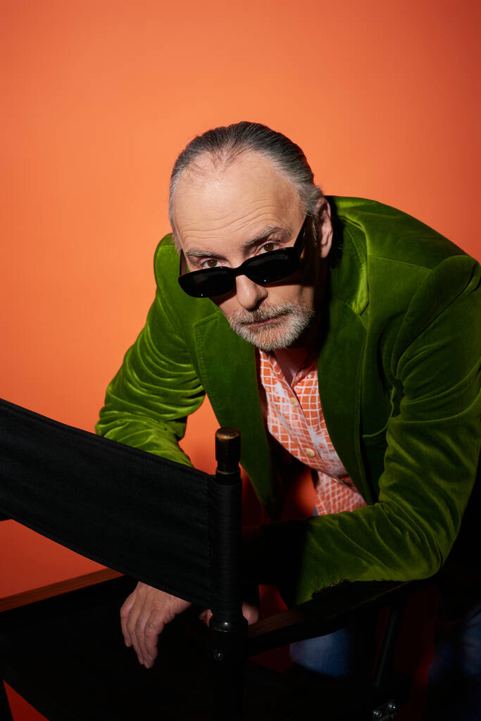 hombre mayor serio y seguro de sí mismo en chaqueta de terciopelo verde mirando a la cámara sobre gafas de sol oscuras y de moda mientras posa cerca de la silla sobre fondo rojo y naranja, concepto de envejecimiento de moda - Foto, Imagen