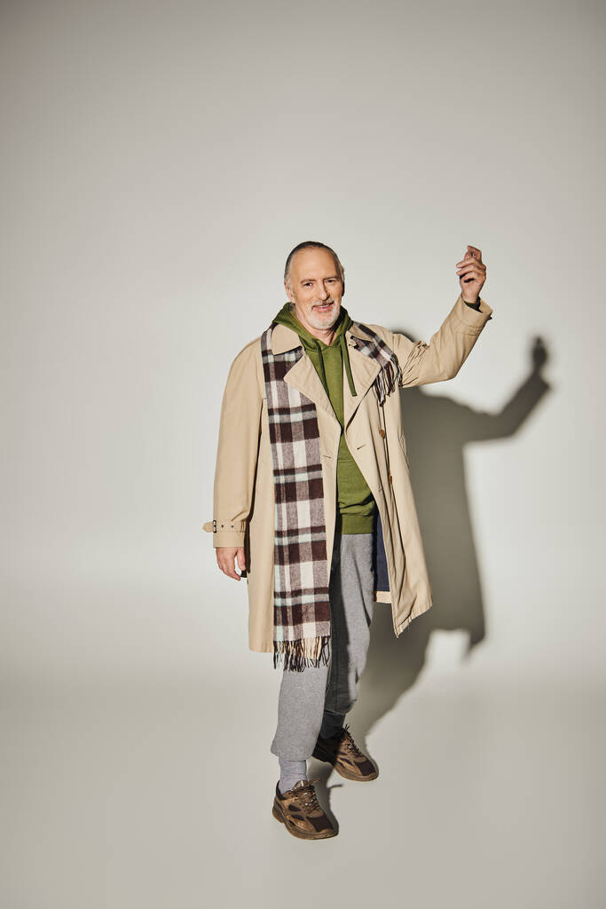 full length of expressive and optimistic senior man in green hoodie, plaid scarf and beige trench coat gesturing and looking at camera on grey background with shadow, happy age concept - Photo, Image