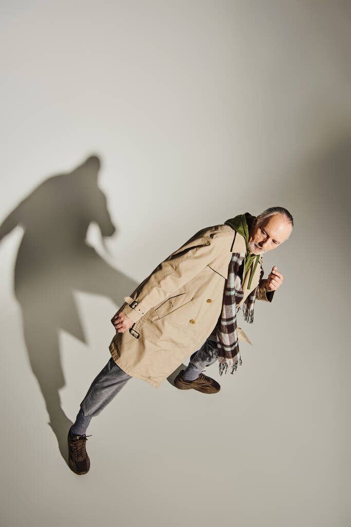 κορυφαία άποψη του μοντέλου με αυτοπεποίθηση ανώτερος σε καρό κασκόλ, πράσινο hoodie και μπεζ παλτό χαρακωμάτων στέκεται σε κομψή στάση σε γκρι φόντο με σκιά, μοντέρνα έννοια γήρανσης - Φωτογραφία, εικόνα