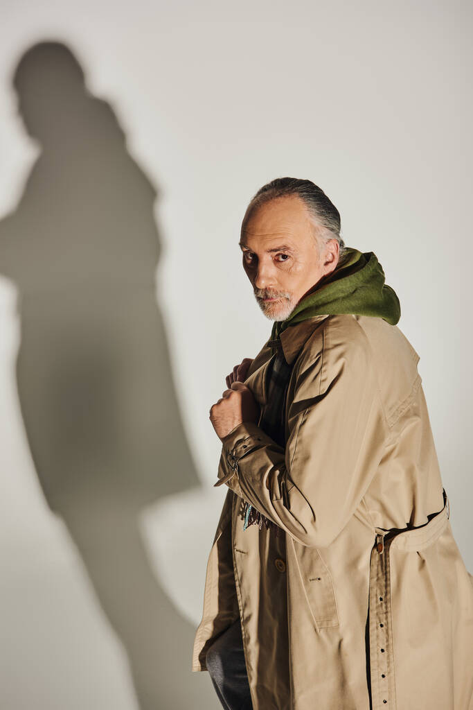 fashion and age, personal style, confident senior man with serious face expression standing in beige trench coat and looking at camera on grey background with shadow - Photo, Image
