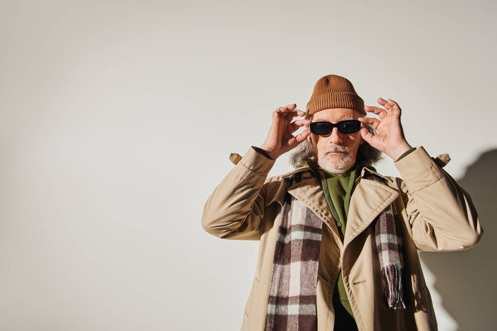 γενειοφόρος ηλικιωμένος άνδρας σε καπέλο beanie, καρό μαντήλι και κομψό παλτό χαρακωμάτων κοιτάζοντας κάμερα, ενώ στέκεται σε γκρι φόντο, hipster στυλ, εκφραστική ατομικότητα, μοντέρνα έννοια γήρανσης - Φωτογραφία, εικόνα