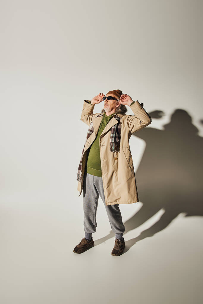 full length of cool senior man in beanie hat and beige trench coat adjusting dark sunglasses and looking away on grey background with shadow, casual hipster style attire, fashion and age concept - Photo, Image