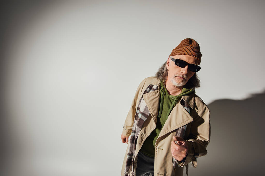 senior and bearded man in dark sunglasses, beanie hat, plaid scarf and beige trench coat looking at camera on grey background with shadow and copy space, hipster style, fashion and age concept - Photo, Image