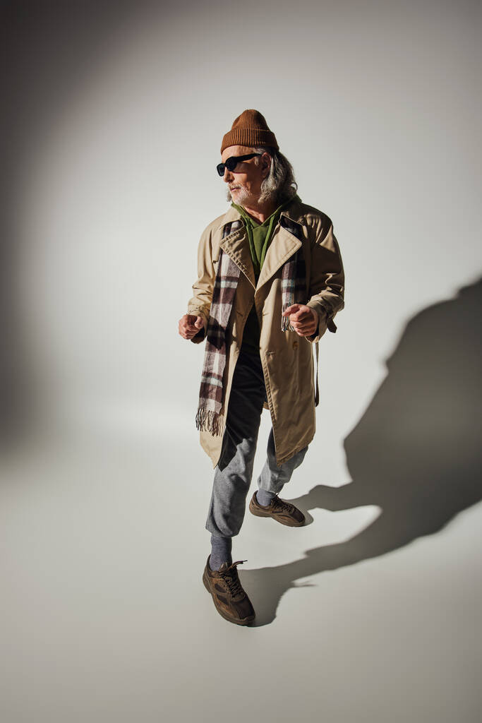 εκφραστική προσωπικότητα, ανώτερος hipster άνθρωπος σε σκούρα γυαλιά ηλίου, καπέλο beanie και μπεζ παλτό χαρακωμάτων στέκεται σε κομψή στάση σε γκρι φόντο με σκιά, θετική και μοντέρνα αντίληψη γήρανσης - Φωτογραφία, εικόνα