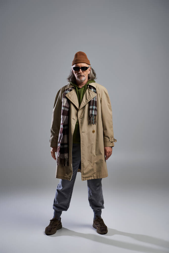 vooraanzicht van trendy en bebaarde senior man met donkere zonnebril, muts, beige trench coat en sneakers op grijze achtergrond, hipster fashion, trendy aging concept, full length - Foto, afbeelding