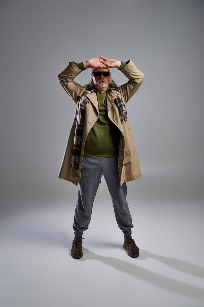 full length of cool and senior hipster man in dark sunglasses, beanie hat, beige trench coat and sneakers posing with hands above head on grey background, fashionable aging concept - Photo, Image