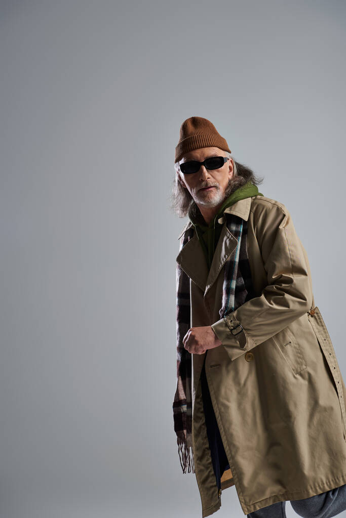 grey haired senior man in dark sunglasses, beanie hat, beige trench coat and plaid scarf looking away on grey background, expressive personality, hipster fashion, positive aging concept - Photo, Image