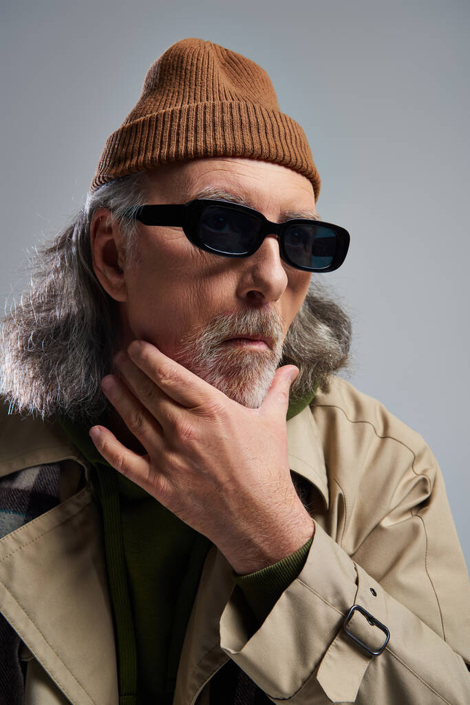 πορτρέτο του στοχαστικού ηλικιωμένου άνδρα με αποθαρρυμένη έκφραση προσώπου, σε καπέλο σκούρα γυαλιά ηλίου και καμπαρντίνα αγγίζοντας γενειάδα σε γκρι φόντο, hipster στυλ, φωτογράφιση μόδας - Φωτογραφία, εικόνα