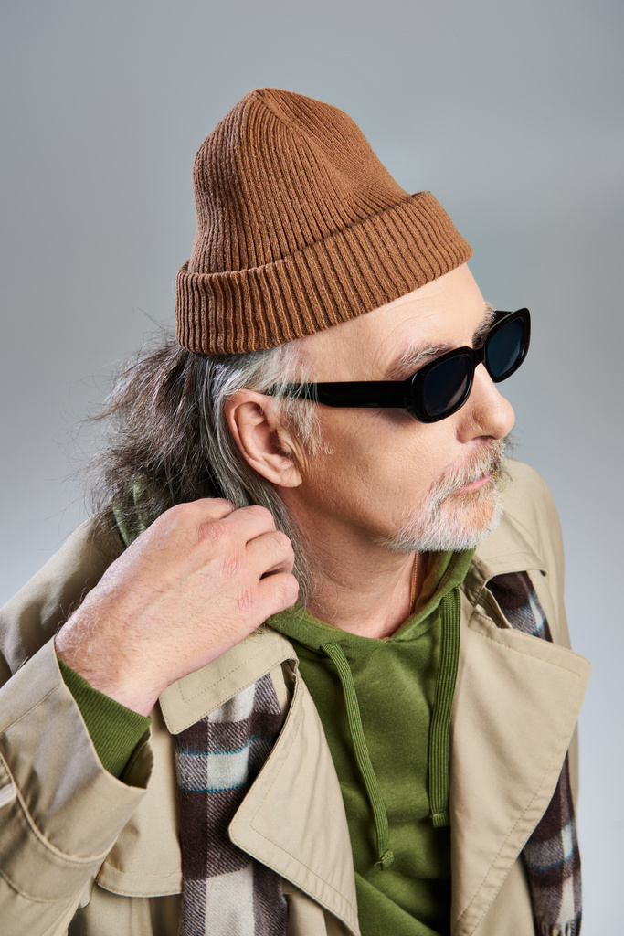 retrato de hombre hipster envejecido en gafas de sol oscuras, gorro sombrero, sudadera con capucha verde y gabardina beige mirando hacia otro lado mientras está de pie sobre fondo gris, envejecimiento con el concepto de estilo, sesión de fotos de moda - Foto, imagen