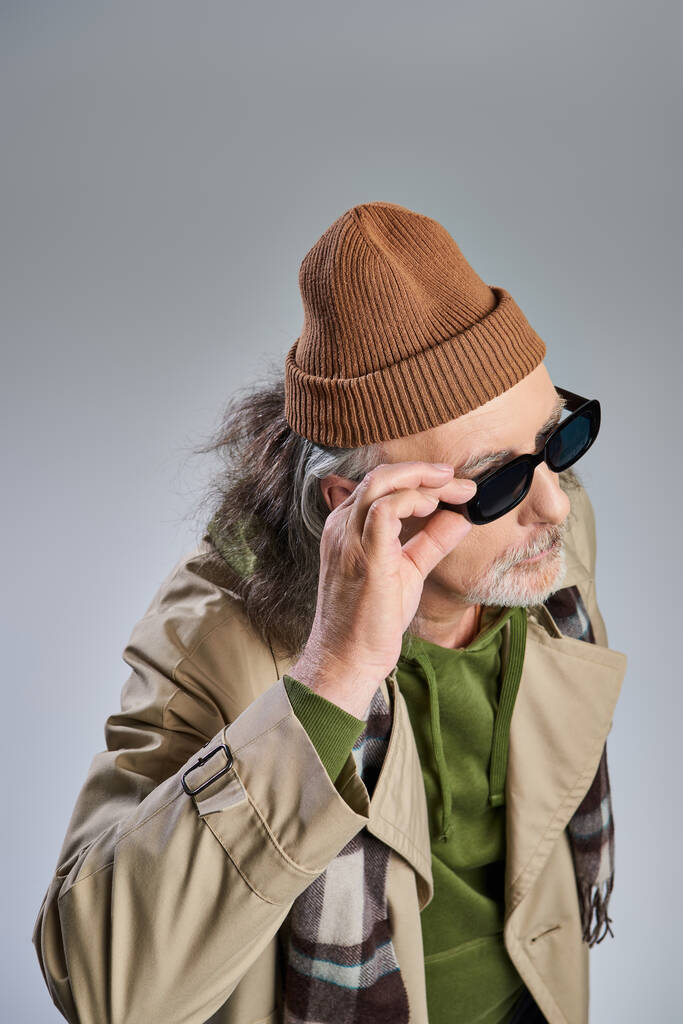 Şapkalı ve bej ceketli şık ve sakallı bir adamın, koyu renk güneş gözlüğü takıp gri arka plana, modaya uygun yaşam tarzına ve yaşlanma konseptine baktığı yüksek açılı görüntüsü. - Fotoğraf, Görsel