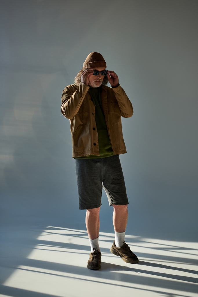 longitud completa de hombre senior de moda ajustando gafas de sol oscuras mientras está de pie en sombrero de gorro, chaqueta y pantalones cortos en fondo gris con iluminación, moda hipster, concepto de envejecimiento positivo y de moda - Foto, Imagen