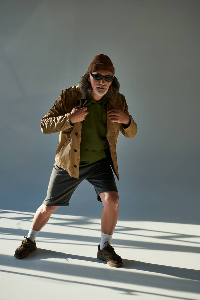 full length of senior and hipster style man in beanie hat, σκούρα γυαλιά ηλίου, σακάκι και σορτς κοιτάζοντας την κάμερα σε γκρι φόντο με φωτισμό, κομψή στάση, μοντέρνα αντίληψη γήρανσης - Φωτογραφία, εικόνα