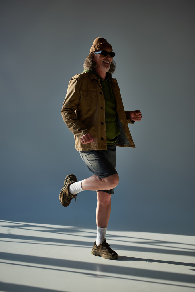 volle Länge von lächelnden und stilvollen männlichen Senior-Modell posiert auf einem Bein auf grauem Hintergrund mit Beleuchtung, Hipster-Mode, dunkle Sonnenbrille, Jacke und Shorts, glücklich und modisch alternden Konzept - Foto, Bild