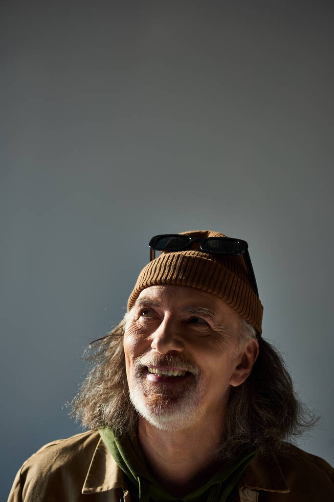 portrait of cheerful and carefree senior man with grey hair and beard, wearing dark sunglasses on beanie hat, looking up on grey background, with copy space, happy and fashionable aging concept - Photo, Image