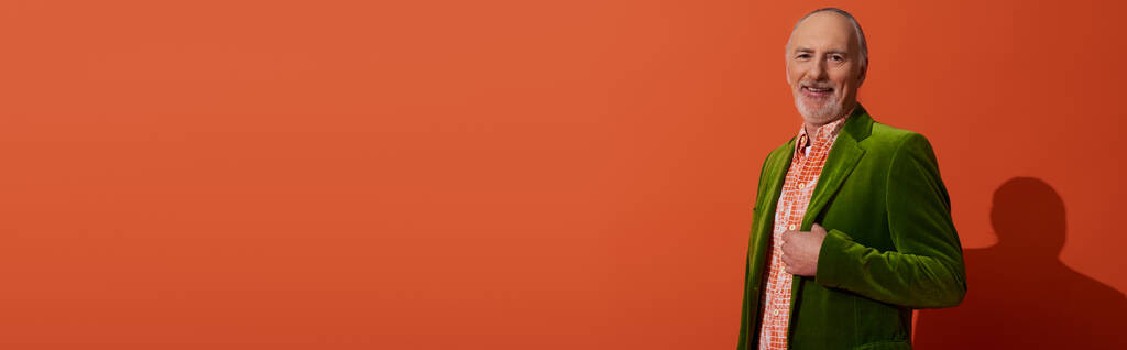 fröhlicher, grauhaariger und bärtiger Mann, der in grünem Velours-Blazer und trendigem Hemd auf rot-orangefarbenem Hintergrund in die Kamera blickt, fröhliches Altern, älterer Stil, Mode- und Alterskonzept, Banner - Foto, Bild