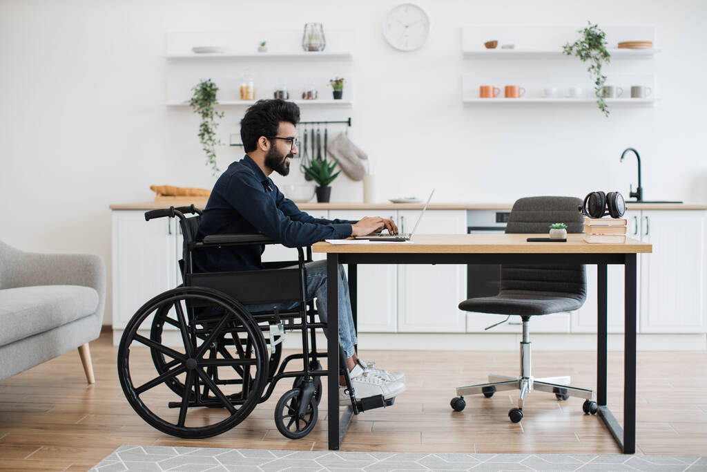 Side view portrait of indian male in wheelchair working at writing desk in open-plan kitchen of modern apartment. Young adult with disability starting working day with checking email account at home. - Photo, Image