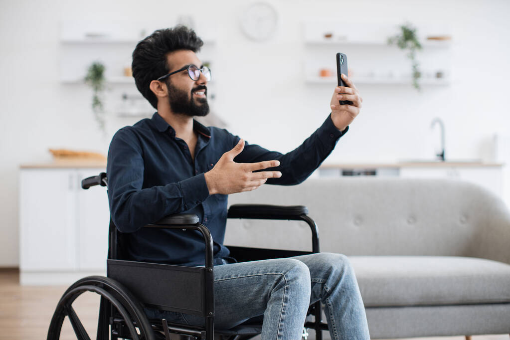 Adulto joven alegre con discapacidad que sostiene móvil mientras planea viajar al aire libre en un apartamento moderno. Sonriente árabe hombre charlando en línea con sus familiares a través de la aplicación de videollamada en el comedor. - Foto, imagen