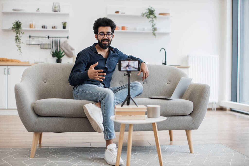 Frontansicht des fröhlichen Content-Machers in Freizeitkleidung, der Videos für Follower auf modernen Smartphones aufzeichnet. Junger indischer Mann mit Brille und Laptop sitzt auf bequemem Sofa im Wohnzimmer. - Foto, Bild