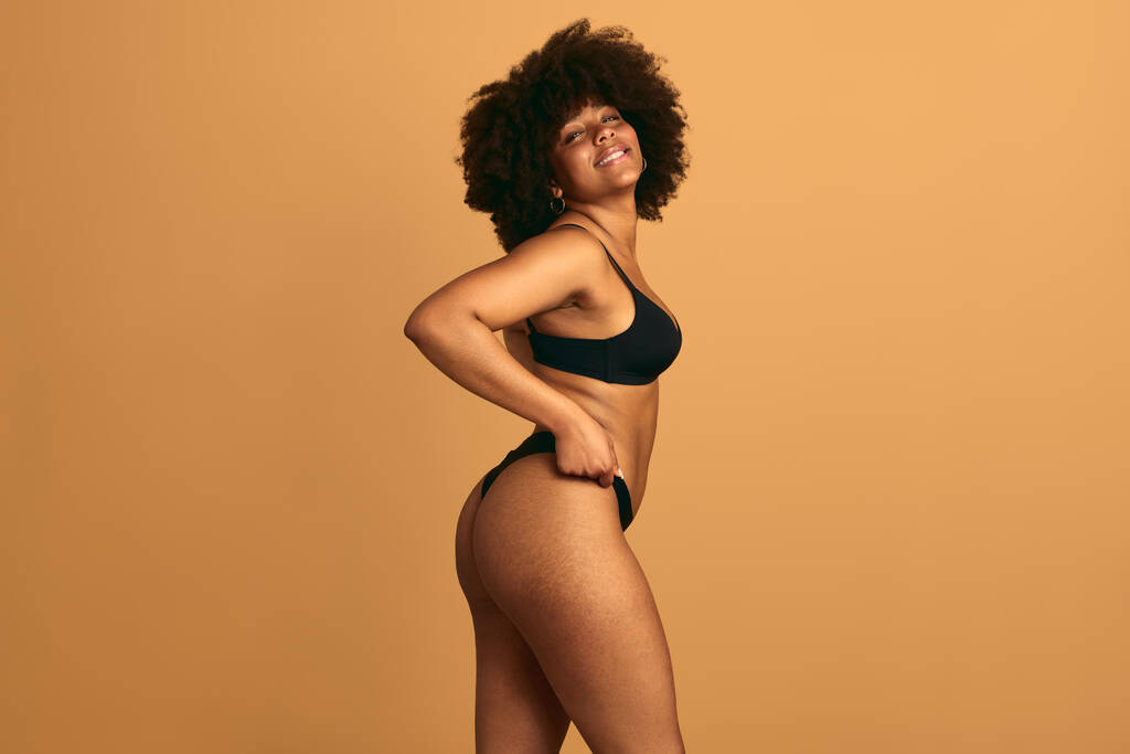 Side view van prachtige jonge Afro-Amerikaanse vrouw model in zwart lingerie met Afro kapsel kijken naar camera en aanraken slipje tegen beige achtergrond - Foto, afbeelding