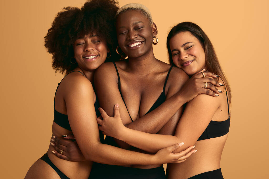Glimlachende zachte multiraciale vriendinnen in zwart ondergoed knuffelen in de studio met ogen gesloten zusterschap en ondersteuning concept - Foto, afbeelding