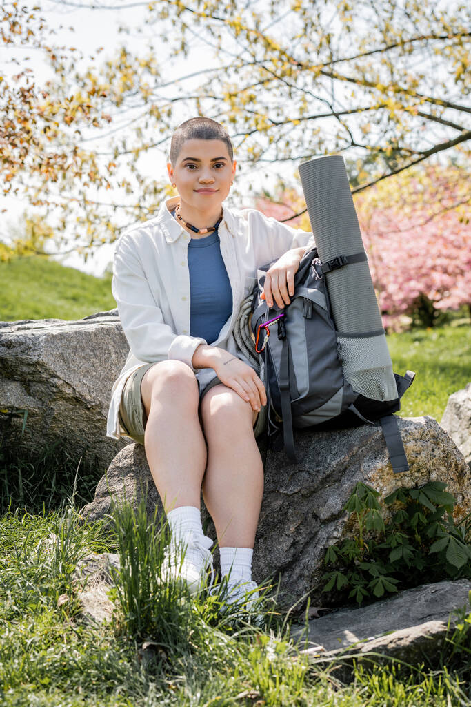 Young short haired and tattooed female traveler looking at camera while sitting near backpack and fitness mat on stone with blurred landscape at background, Translation of tattoo: love - Photo, Image