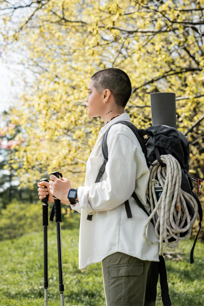 Side view of young short haired and tattooed female hiker with backpack and climbing rope holding trekking poles and walking on blurred nature, independent traveler embarking on solo journey - Photo, Image