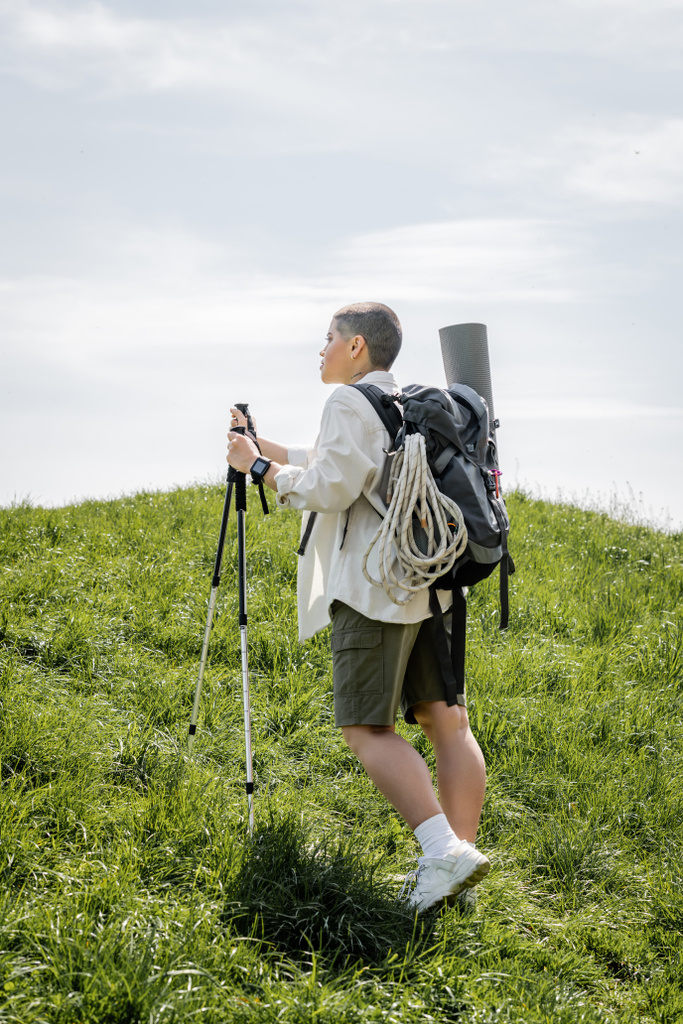 Boční pohled na mladou krátkosrstou cestovatelku s batohem a horolezeckým lanem procházející se s trekkingovými tyčemi na kopci s trávou v pozadí, nezávislá cestovatelka vyrážející na sólovou cestu - Fotografie, Obrázek