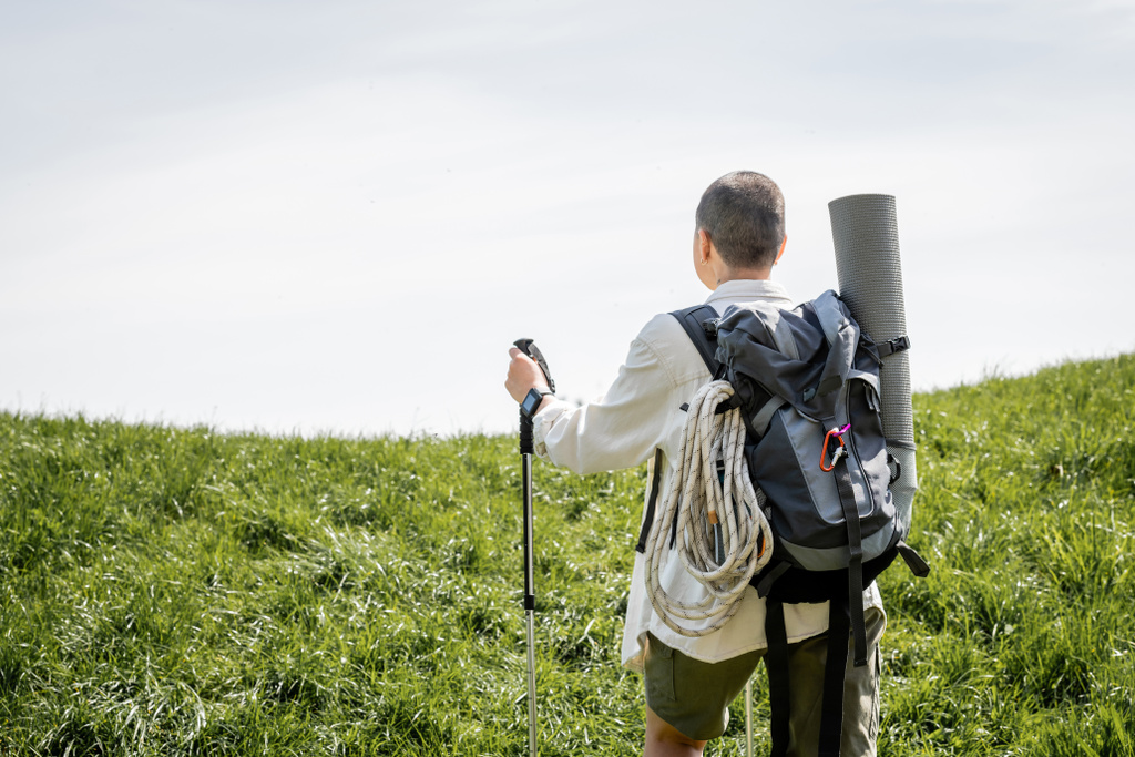 Back view of young short haired woman traveler with backpack holding trekking pole while walking on grassy hill at background, independent traveler embarking on solo journey - Photo, Image