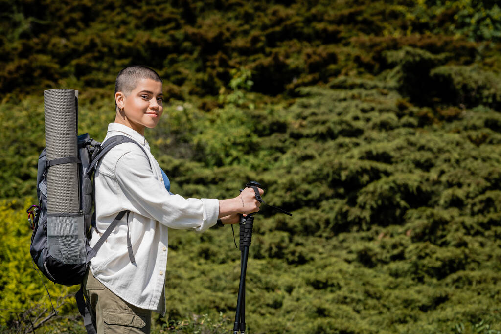 Sonriente joven excursionista de pelo corto con mochila sosteniendo bastones de trekking y mirando a la cámara con la colina y el paisaje en el fondo, Traducción del tatuaje: amor - Foto, imagen