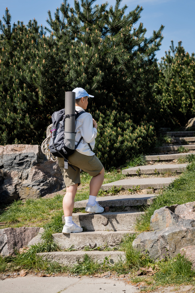 Junge Wanderin in Baseballkappe und lässiger Kleidung mit Rucksack und Reiseausrüstung auf Treppen mit Natur und Himmel im Hintergrund, neugierige Wanderin erkundet neue Landschaften - Foto, Bild