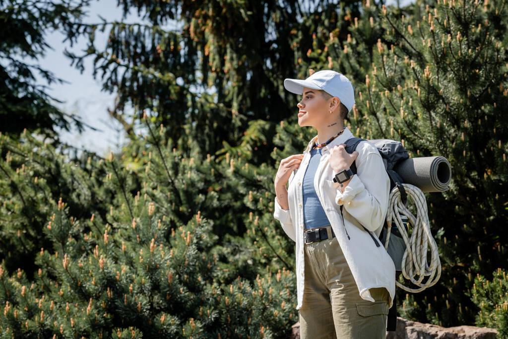 Vista lateral de una joven turista de pelo corto con ropa casual y gorra de béisbol mirando hacia otro lado mientras está de pie con árboles borrosos al fondo, curiosa excursionista explorando nuevos paisajes - Foto, Imagen