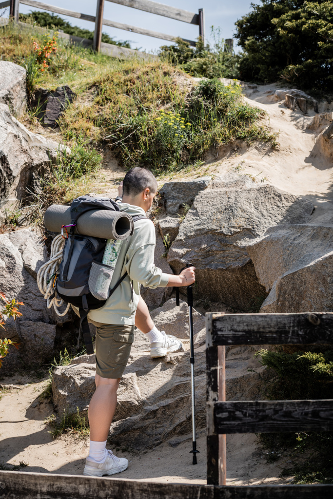 Mladá krátkosrstá turistka s batohem a trekkingovou tyčí kráčející po kopci s kameny a přírodou v pozadí v létě, žena na túrách po rozlehlé krajině, Překlad tetování: láska - Fotografie, Obrázek