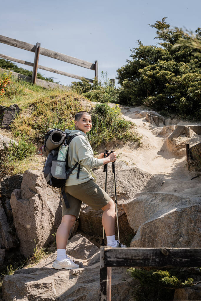 Joven viajero femenino de pelo corto positivo con mochila sosteniendo bastones de trekking y de pie cerca de la colina con piedras al fondo durante el verano, Traducción del tatuaje: amor - Foto, imagen