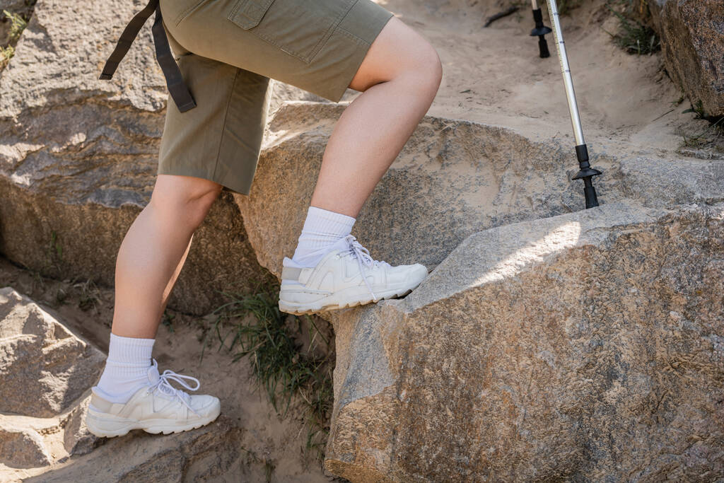 Cropped view of young woman in casual clothes walking near trekking poles on stones on hill, tranquil hiker finding inner peace on trail, summer, activity, fresh air  - Photo, Image