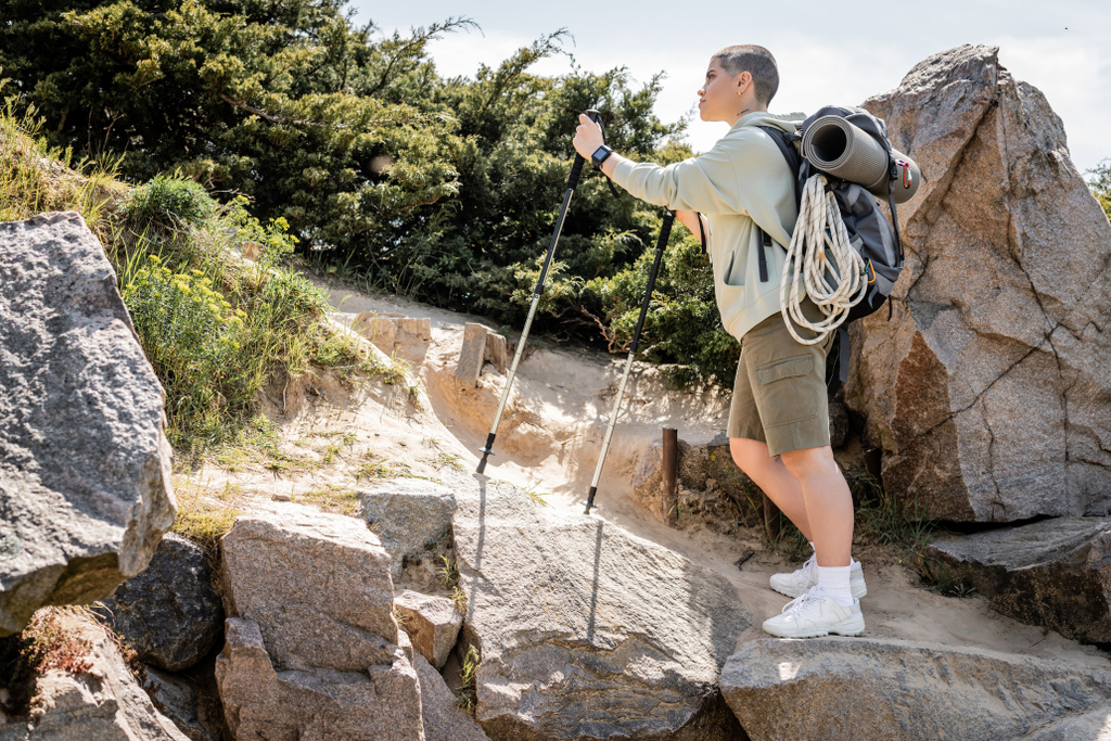 Πλάγια άποψη της νεαρής κοντή γυναίκα τουρίστρια με σακίδιο κρατώντας περιπατητικούς πόλους περπάτημα στο λόφο με πέτρες και γρασίδι στο παρασκήνιο, ήσυχο πεζοπόρος εύρεση εσωτερική ειρήνη στο μονοπάτι, το καλοκαίρι - Φωτογραφία, εικόνα