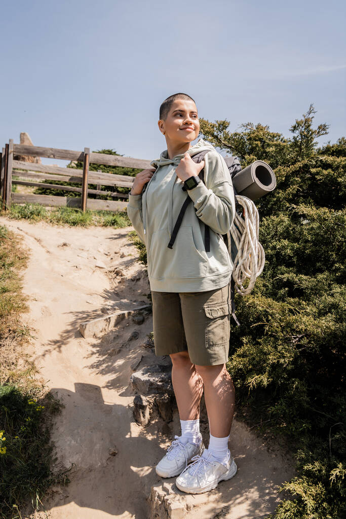 Usmívající se a krátkosrstá turistka s batohem, fitness rohožkou a lezeckým lanem stojící a dívající se na kopec s oblohou na pozadí, turistka nacházející inspiraci v přírodě - Fotografie, Obrázek