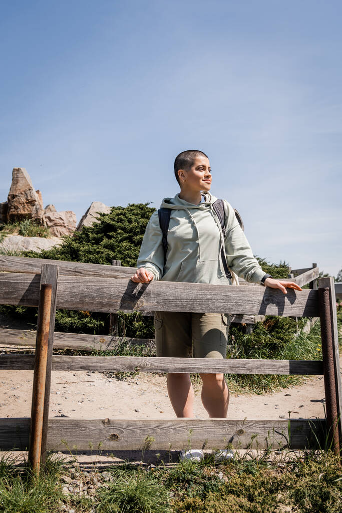 Smiling young short haired female traveler with backpack in casual clothes looking away while standing near wooden fence with nature at background, hiker trekking through landscape, summer - Photo, Image